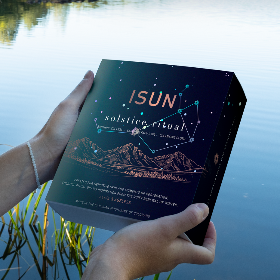 A person standing by a lake holds a box titled 'ISUN: solstice ritual' in both hands, with the cover featuring a dark starry sky and mountain illustration.