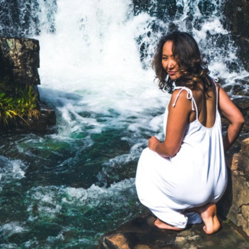 A woman in a white sleeveless dress crouches on a rock beside a rushing waterfall in a forest setting.