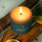 Candle in a container labeled 'Journey of Light' on a wooden surface with an orange slice and plant in the background.