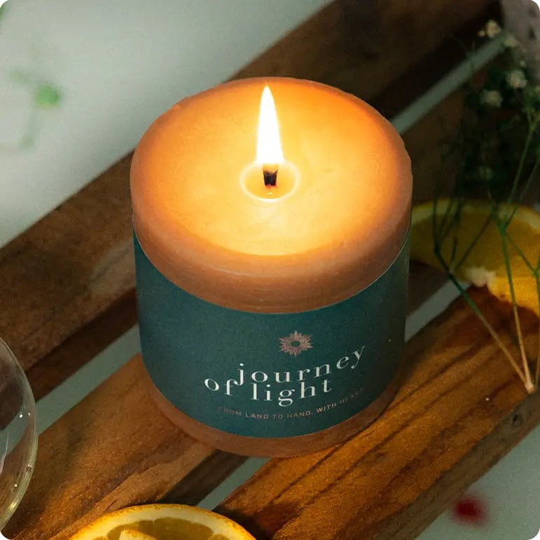 Candle in a container labeled 'Journey of Light' on a wooden surface with an orange slice and plant in the background.