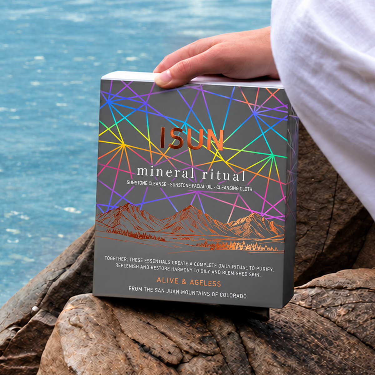A person in white pants sits on rocks by the water, holding a box titled 'ISUN Mineral Ritual' with a teal cover featuring geometric line art.