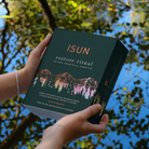 A woman in a green top holds a book titled 'ISUN restore ritual' outdoors by a reflective pond with green foliage and blue water in the background.