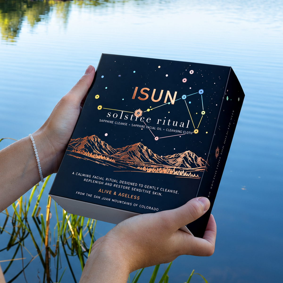 A person standing by a lake holds a box titled 'ISUN: solstice ritual' in both hands, with the cover featuring a dark starry sky and mountain illustration.