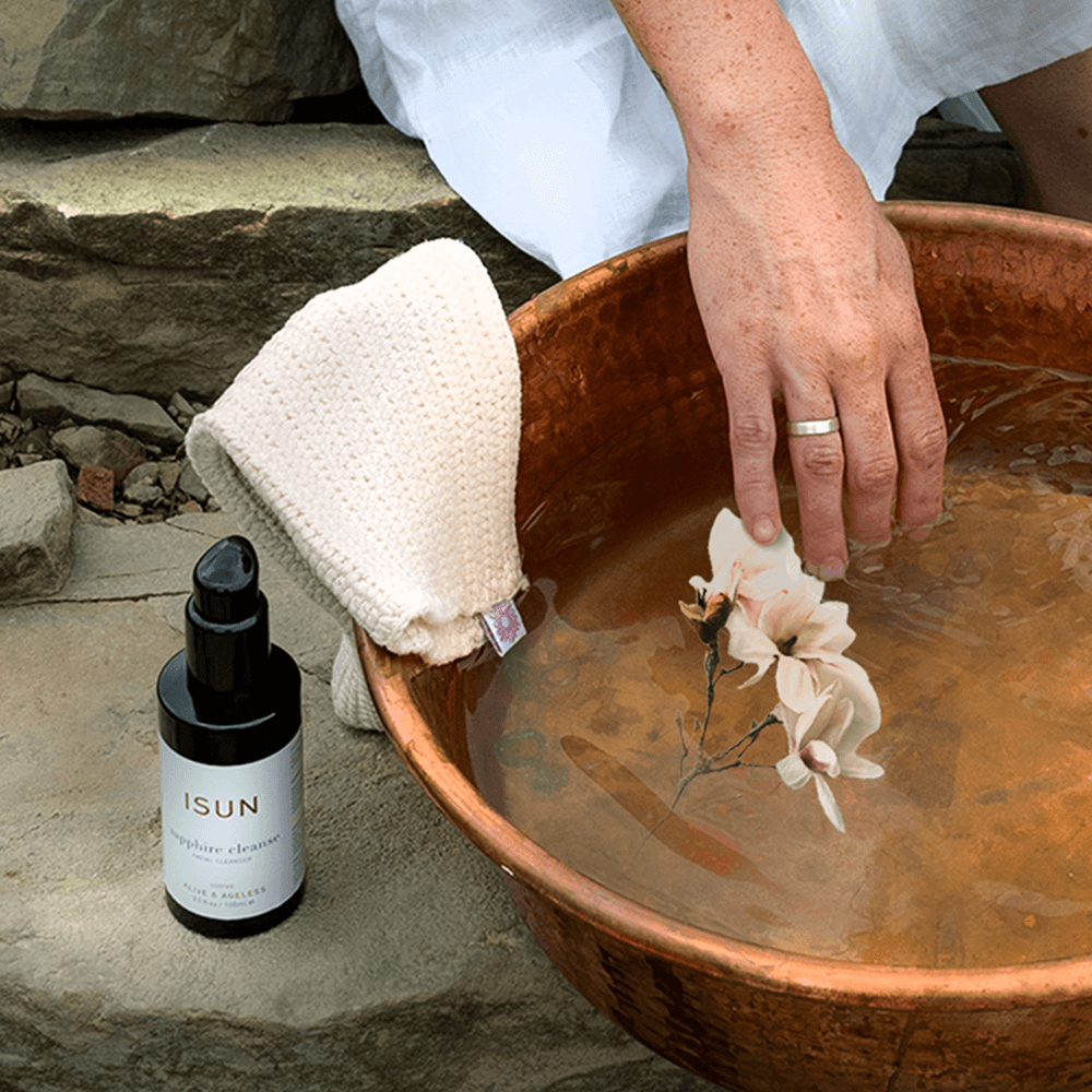 Person dipping a flower into a copper basin with a bottle of ISUN skincare product and cleansing cloth nearby.