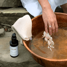 Person dipping a flower into a copper basin with a bottle of ISUN skincare product and cleansing cloth nearby.