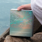A hand holds a square skincare box labeled 'ISUN mineral ritual' outdoors near water and rocks. The box has a muted teal cover with geometric line art and peach mountain graphics.