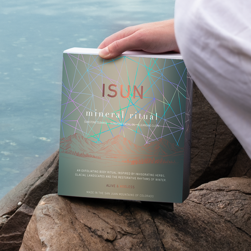 A person in white pants sits on rocks by the water, holding a box titled 'ISUN Mineral Ritual' with a teal cover featuring geometric line art.