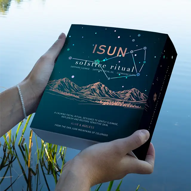 A person holds a dark blue box labeled ISUN solstice ritual in front of a blurred outdoor water scene. The box features a starry night sky, constellation lines, mountain illustrations, and small gold text.