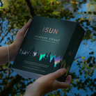 A woman in a green top holds a book titled 'ISUN restore ritual' outdoors by a reflective pond with green foliage and blue water in the background.